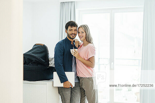 Smiling male freelancer with young woman standing by baby carrier in bright living room