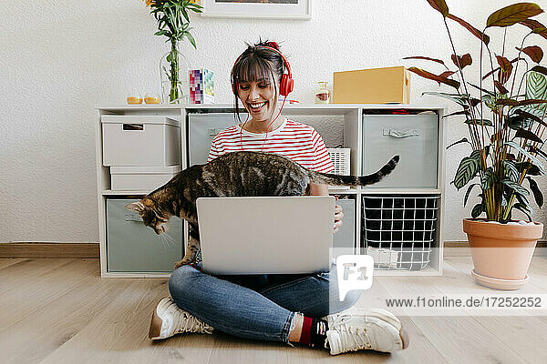 Junge Frau mit Kopfhörern sitzt mit Laptop und Katze zu Hause