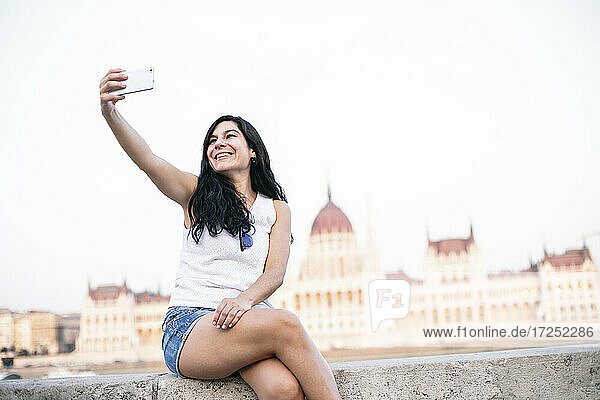 Beautiful woman taking selfie through mobile phone while sitting on retaining wall  Budapest  Hungary