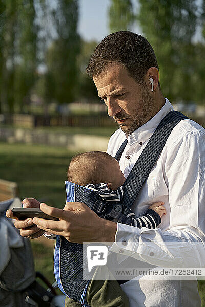 Man with in-ear headphones and baby boy using mobile phone at park