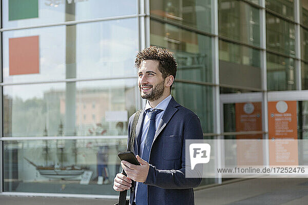Smiling male professional holding shoulder bag and mobile phone during sunny day