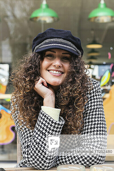 Smiling curly hair woman in cap sitting with hand on chin at sidewalk cafe