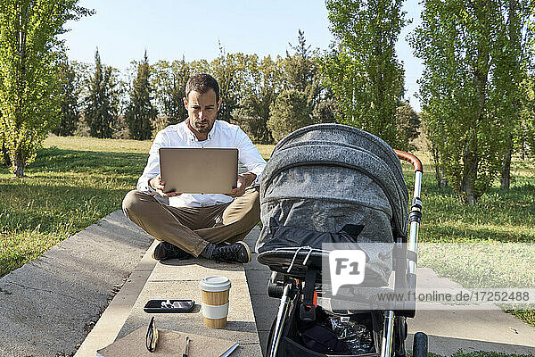 Male entrepreneur using laptop while sitting cross-legged by baby stroller at park