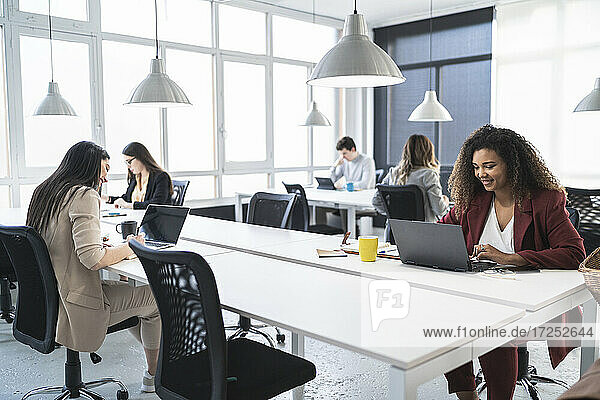 Unternehmerinnen lächelnd bei der Verwendung eines Laptops mit Kollegen im Hintergrund im Büro