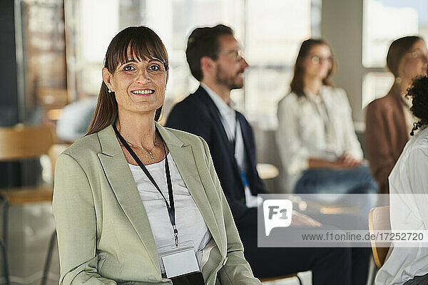Lächelnde Geschäftsfrau sitzt mit Kollegen bei einer Bildungsveranstaltung im Büro