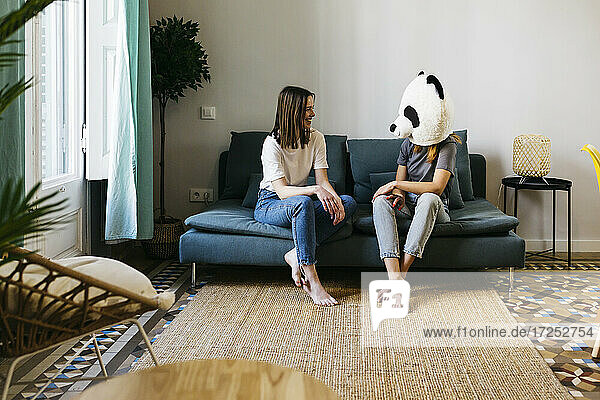 Lächelnde Frau sieht ihre Freundin an  die im Wohnzimmer eine Panda-Maske trägt