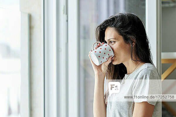 Frau schaut durch das Fenster  während sie zu Hause Kaffee trinkt