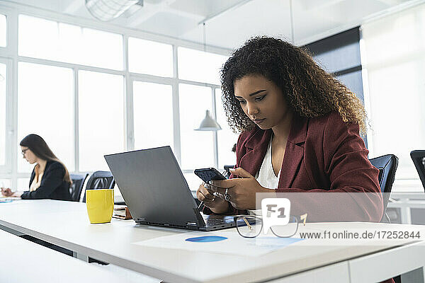 Geschäftsfrau  die ein Mobiltelefon benutzt  während im Hintergrund eine Kollegin in einem Coworking-Büro arbeitet
