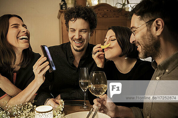 Fröhliche Frau zeigt Smartphone an männliche und weibliche Freunde bei Geburtstagsfeier zu Hause