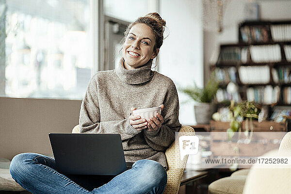 Businesswoman with laptop smiling while holding bowl at coffee shop