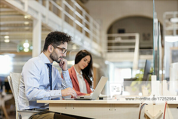 Male professional with hand on chin working on laptop with female colleague in background at coworking office