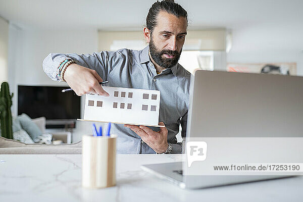 Männlicher Architekt  der ein architektonisches Modell hält  während er zu Hause einen Videoanruf tätigt