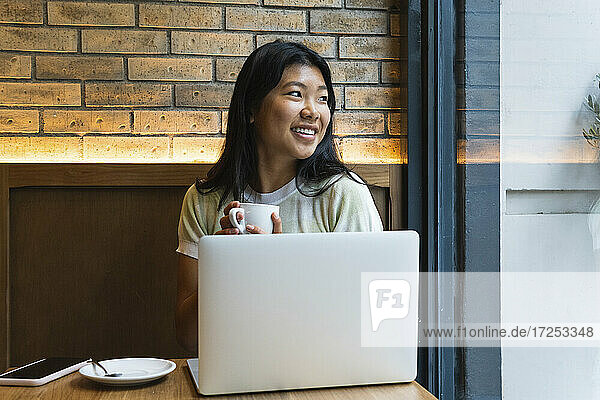 Junge Frau mit Laptop schaut weg und hält eine Kaffeetasse in einem Café
