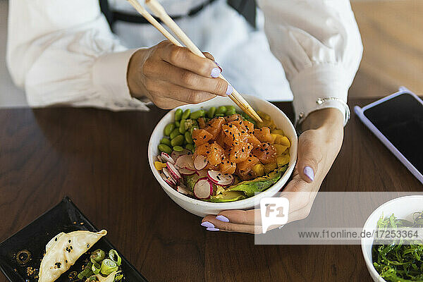 Young woman eating poke bowl with chopsticks on table at restaurant