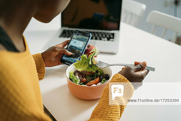 Junge Frau frühstückt gesund  während sie zu Hause ihr Smartphone benutzt