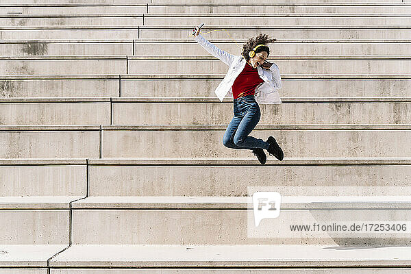 Ecstatic woman with headphones and smart phone jumping on staircase during sunny day