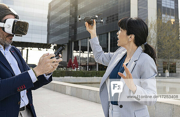 Businessman wearing virtual reality headset operating drone remote while standing with female colleague in office park