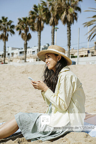 Lächelnde Frau schaut weg  während sie ihr Smartphone am Strand an einem sonnigen Tag hält