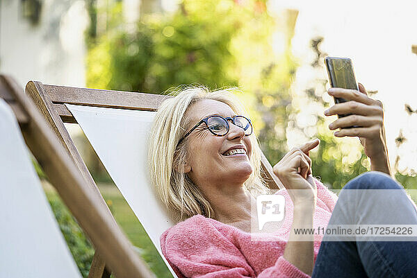 Lächelnde reife Frau  die ein Smartphone benutzt  während sie auf einem Liegestuhl sitzt