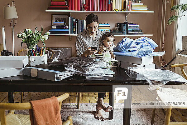 Arbeitende Mutter mit Smartphone beim Sitzen mit Baby Junge im Büro zu Hause