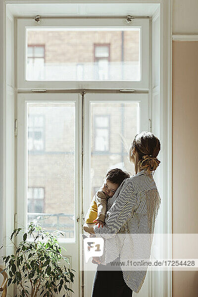 Mutter trägt männliches Kleinkind beim Blick durch das Fenster zu Hause