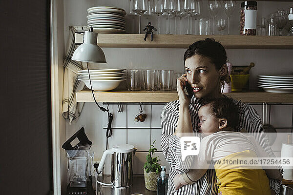 Mutter spricht am Smartphone  während sie ein männliches Kleinkind in der Küche trägt