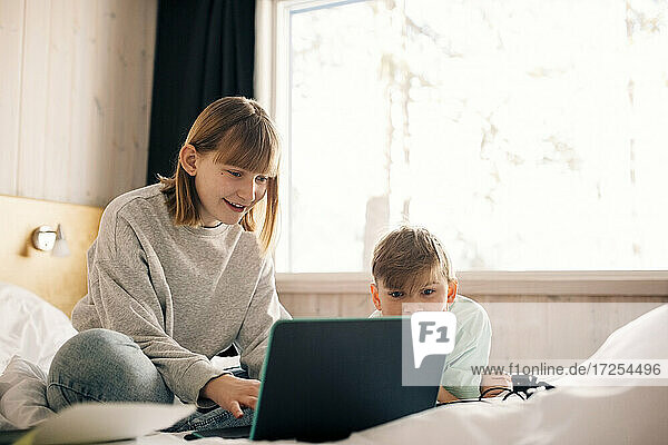Smiling girl with brother using laptop in bedroom at home