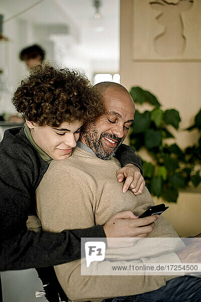 Lächelnder Junge  der seinen Vater von hinten umarmt  während er ein Smartphone benutzt