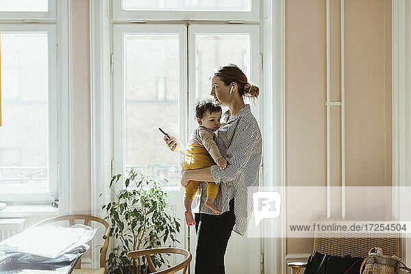 Geschäftsfrau trägt Baby Junge während hält Smartphone zu Hause
