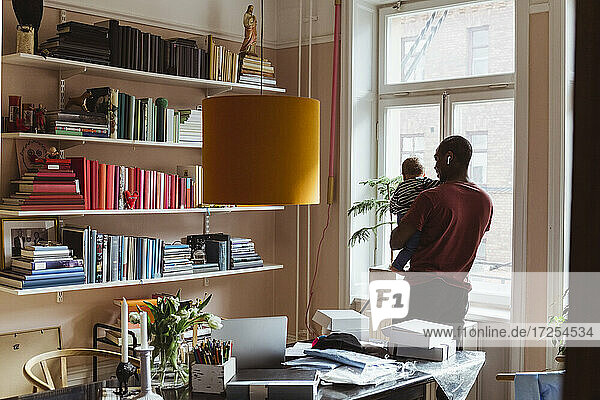 Männlicher Unternehmer trägt Kleinkind im Stehen im Home Office