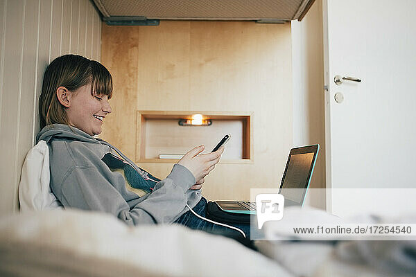 Lächelnde Mädchen Text-Messaging durch Handy  während auf dem Bett im Urlaub zu Hause sitzen