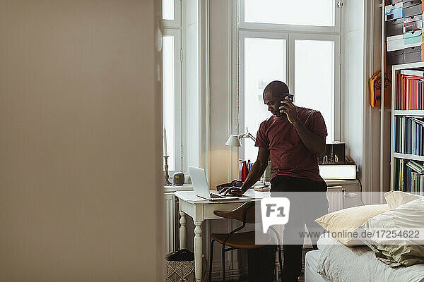 Männlicher Berufstätiger  der einen Laptop benutzt  während er zu Hause mit einem Smartphone telefoniert