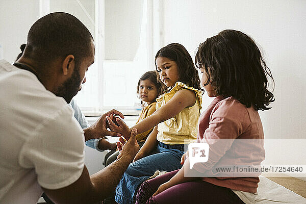 Male healthcare worker examining girl's hand sitting by sisters at medical clinic