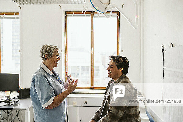 Älterer weiblicher Arzt gestikuliert  während er mit einem jungen männlichen Patienten in einer medizinischen Klinik diskutiert