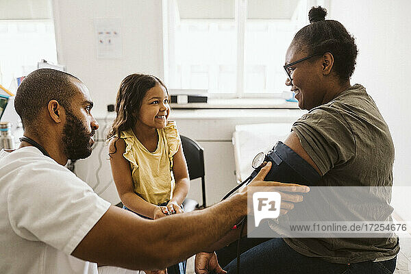 Lächelnde Mutter und Tochter schauen sich gegenseitig an  während ein männlicher Arzt den Blutdruck misst