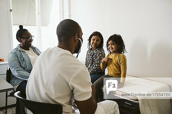 Lächelndes Mädchen  das bei seiner Schwester sitzt und gestikuliert  während es einen männlichen Arzt in einer medizinischen Klinik ansieht