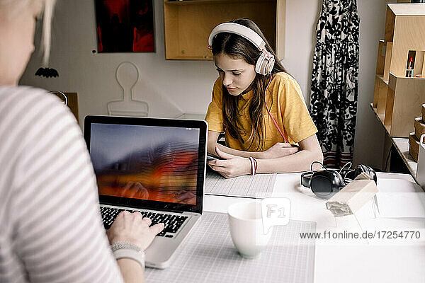 Girl wearing headphone E-learning through digital tablet while sitting with businesswoman working on laptop at desk
