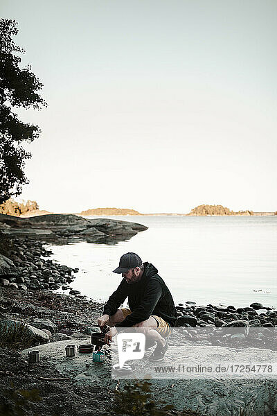 Mann in voller Länge beim Kochen von Essen  während er auf einem Felsen am Seeufer hockt
