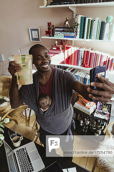 Lächelnder Geschäftsmann nimmt Selfie mit Paket und Sohn in Träger zu Hause