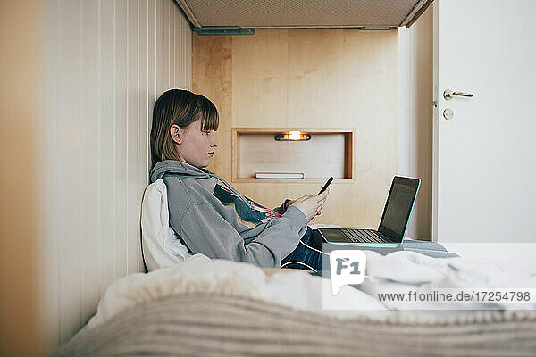 Teenager-Mädchen sitzt mit Laptop mit Smartphone in Ferienhaus