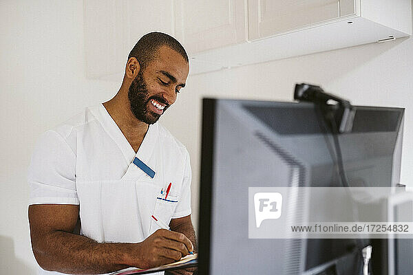 Smiling male healthcare worker writing while consulting patient through video call
