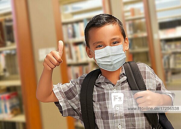Hispanic student boy wearing face mask with thumbs up and backpack in the library