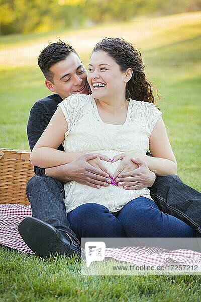 Pregnant hispanic couple making heart shape with hands on belly in the park outdoors