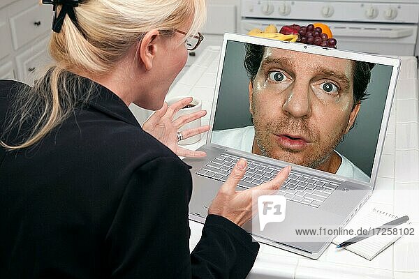 Shocked woman in kitchen using laptop with strange man on screen