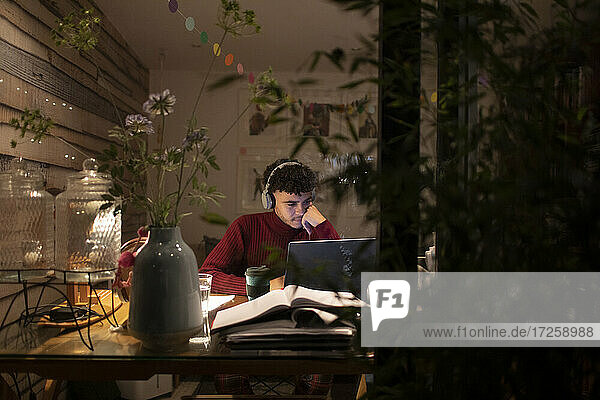 Konzentrierter junger Mann mit Kopfhörern arbeitet spät am Laptop im Heimbüro