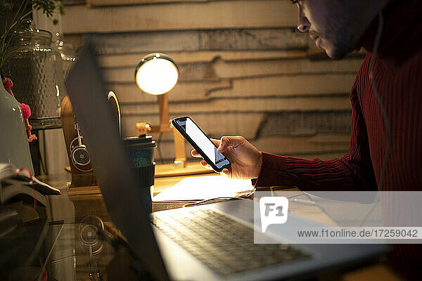 Junger Mann mit Smartphone am Laptop im Home-Office