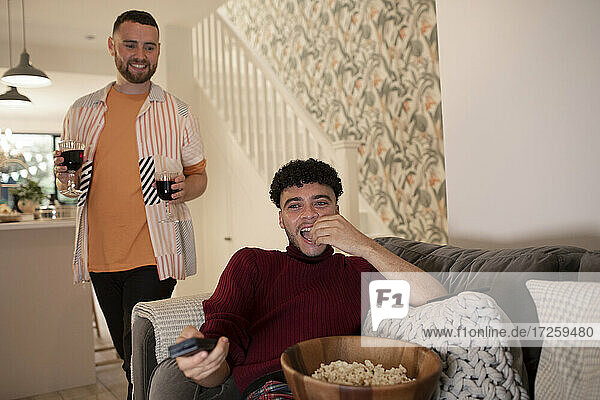 Glückliches schwules männliches Paar mit Rotwein und Popcorn beim Fernsehen zu Hause