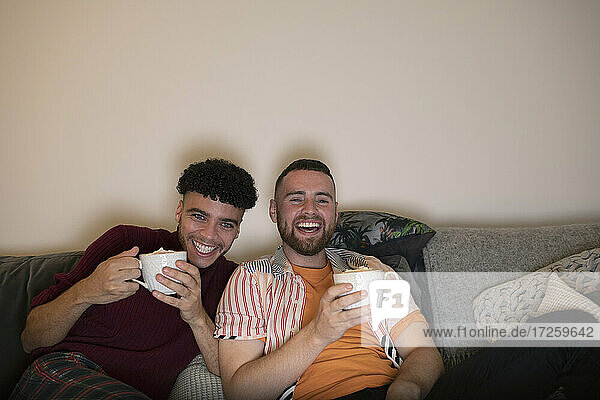Happy gay male couple drinking hot cocoa and watching TV on sofa