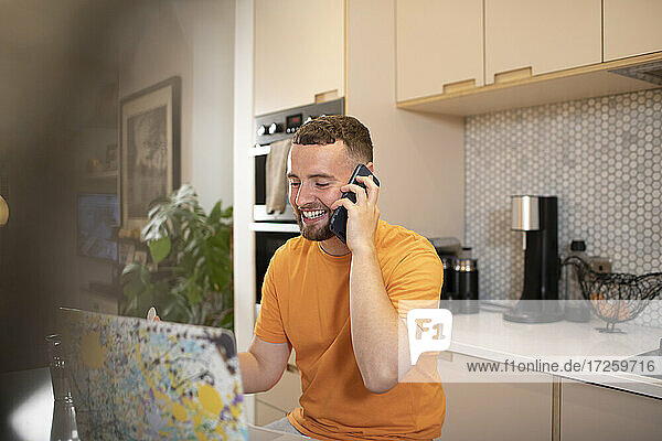 Happy young man working from home talking on smart phone at laptop