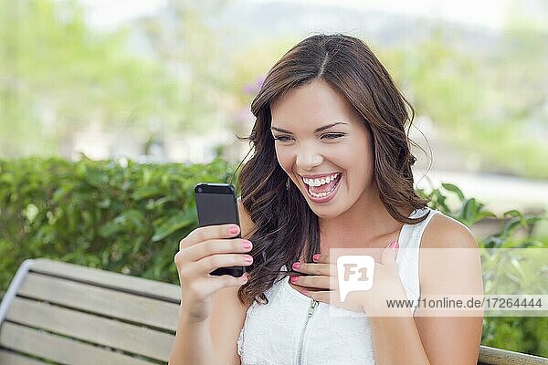 Attractive smiling young adult female texting on cell phone outdoors on a bench